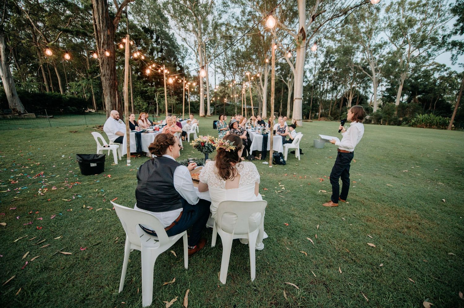 Outdoor wedding reception under string lights and tall trees at dusk with guests seated at tables on a lush lawn.