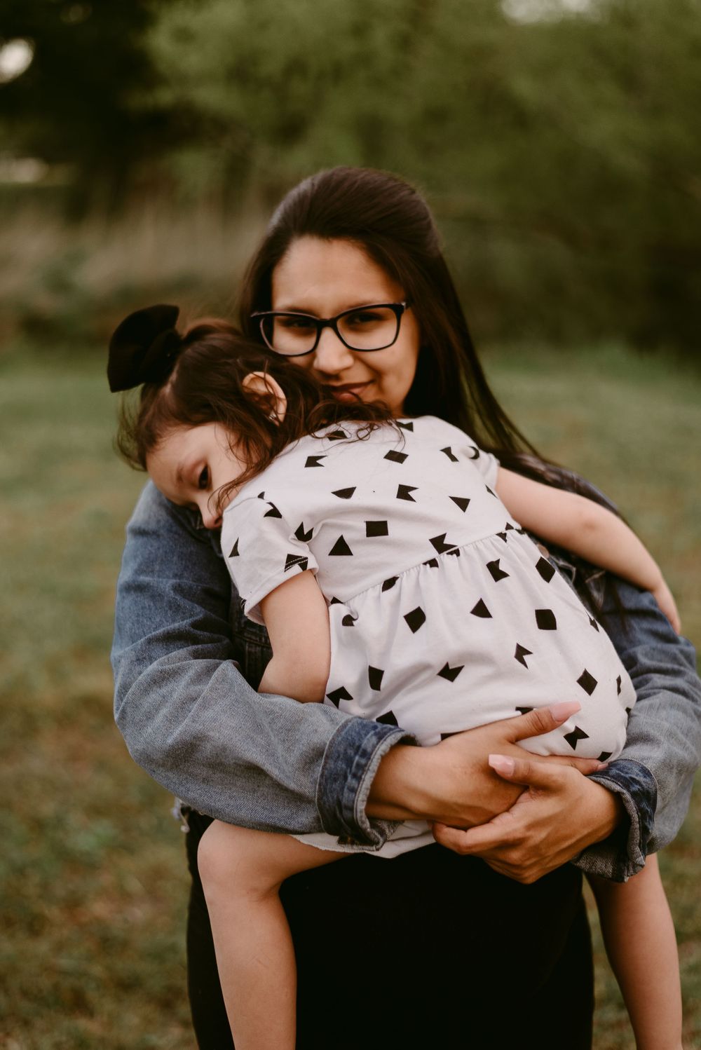 A tender embrace captured outdoors showing a denim jacket and white patterned dress in warm natural lighting.