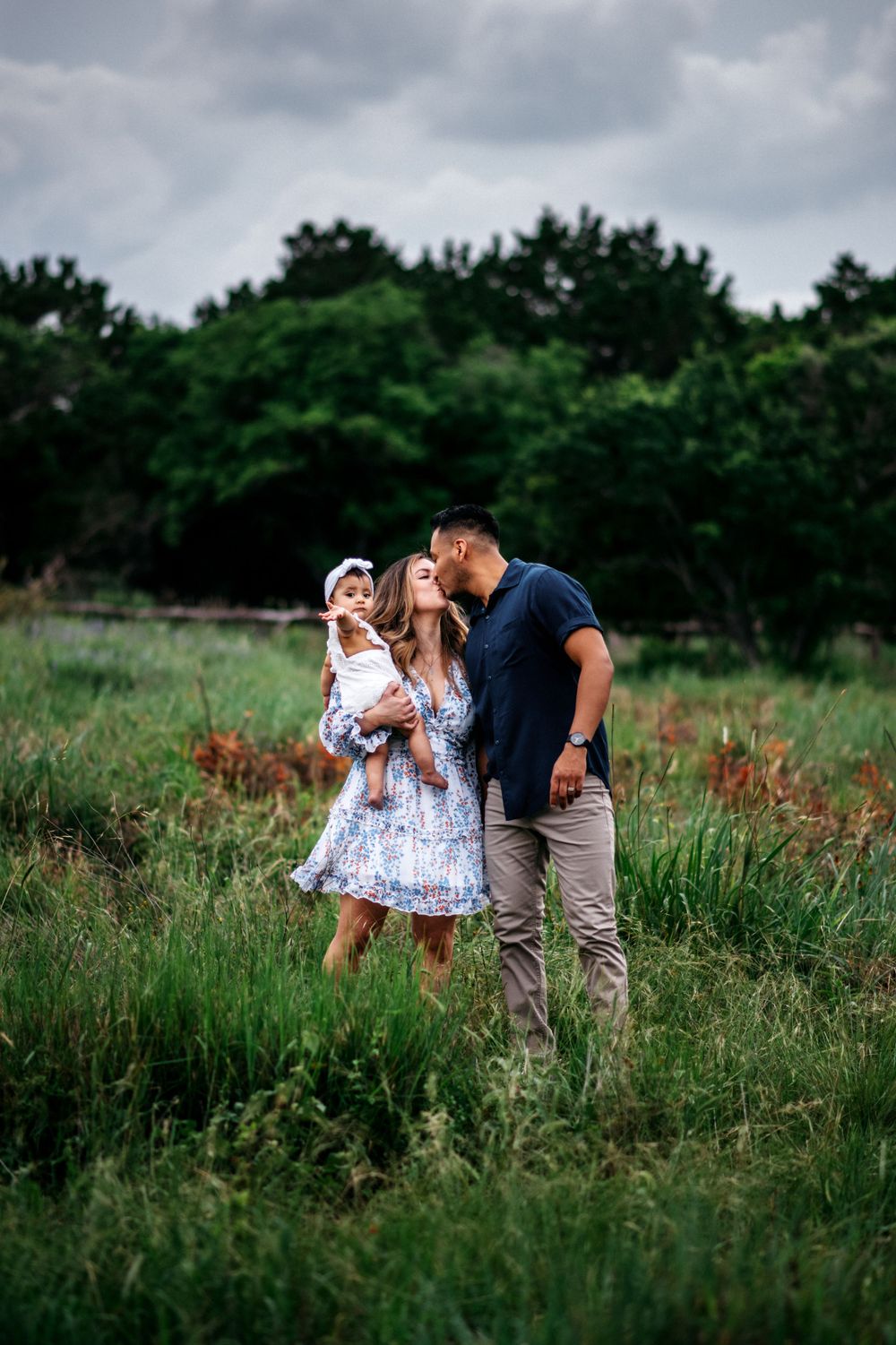 Mother and father kissing while holding their one year old in a field in San Antonio, Texas.