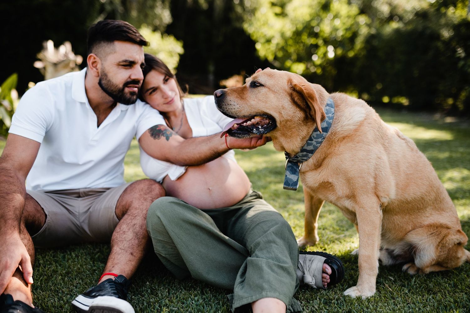 A couple lounges on grass with their friendly dog joining them outdoors.