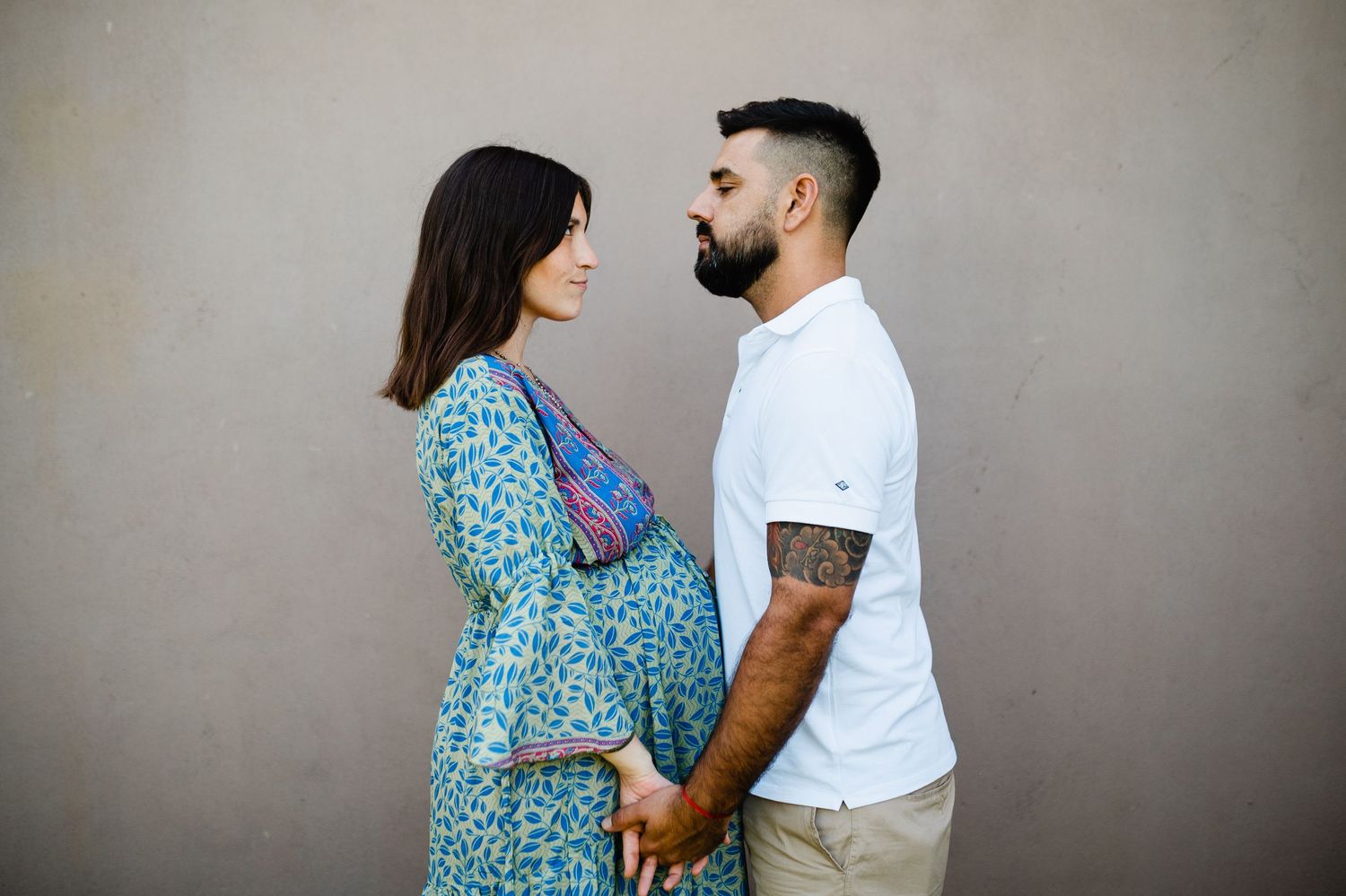 Couple in casual attire embraces against a neutral wall during a photo session.