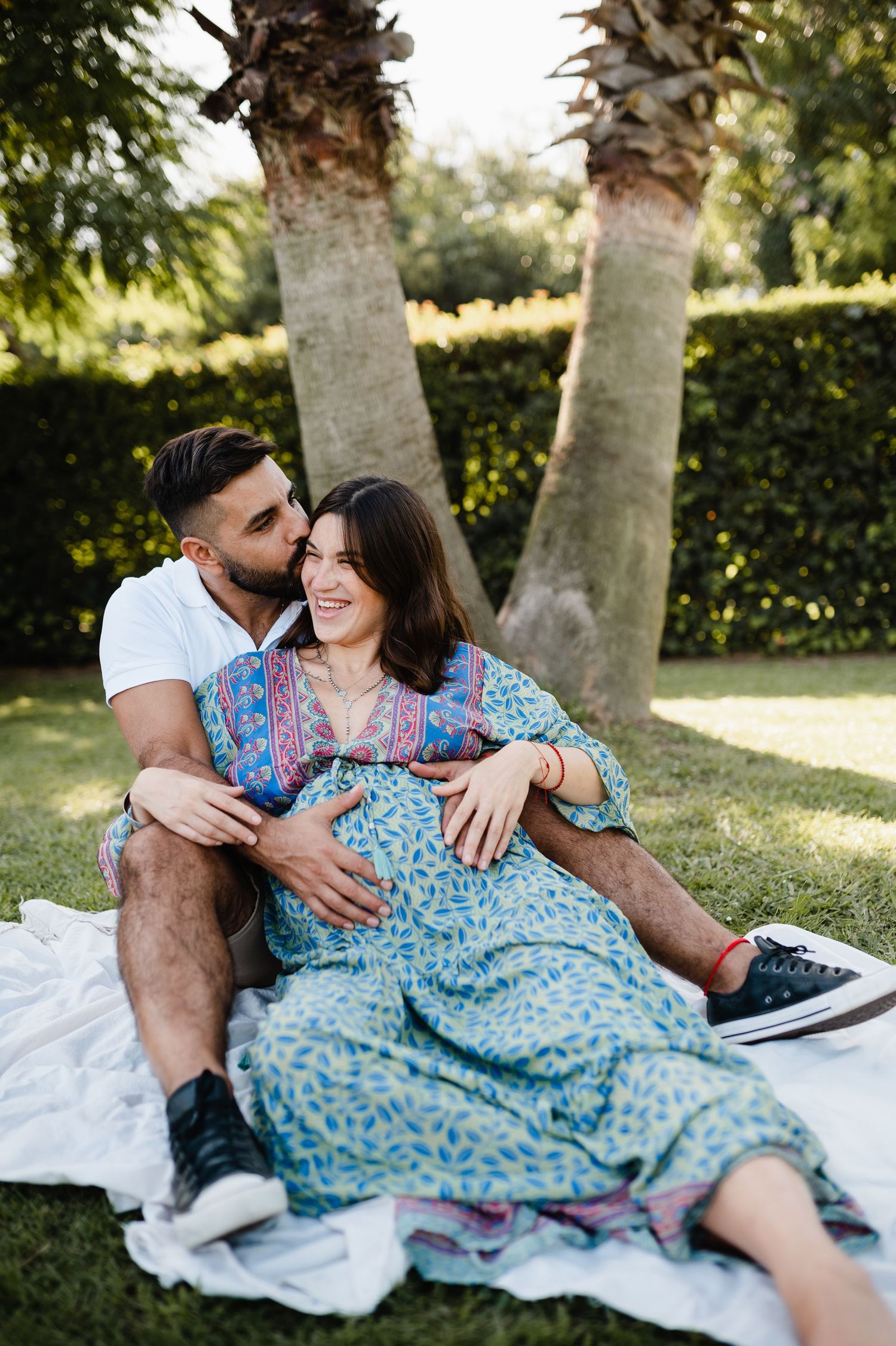 Intimate couple sharing a tender moment while sitting on blanket under palm trees in garden setting.