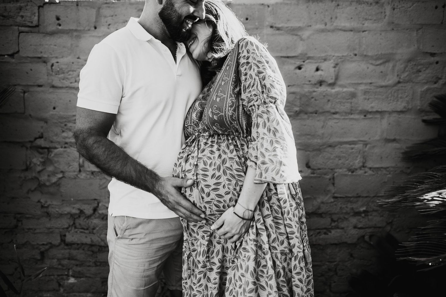 Couple in coordinating outfits embrace against brick wall during maternity photoshoot.
