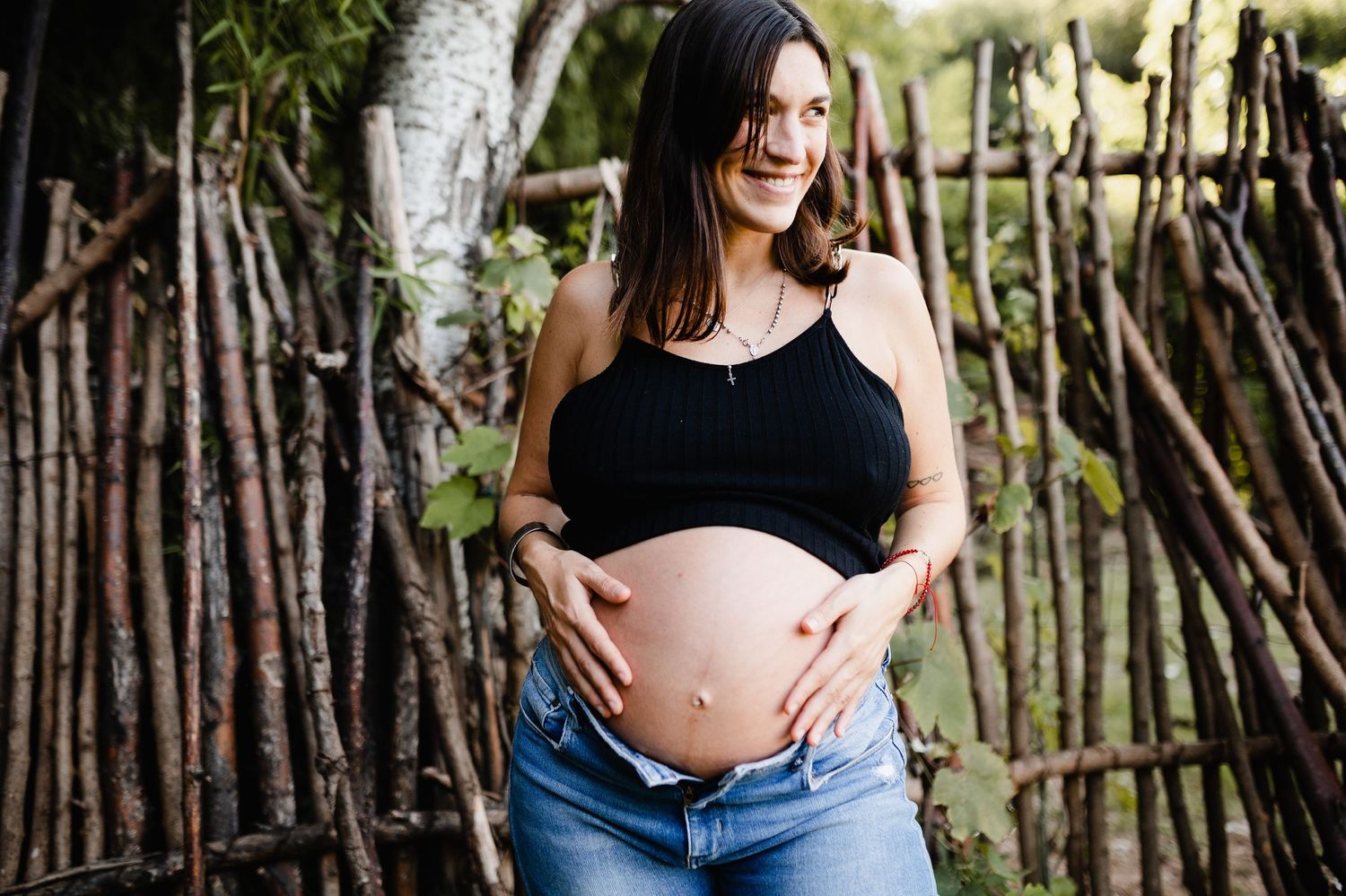 Expectant mother in black top and jeans poses against bamboo backdrop in garden setting.