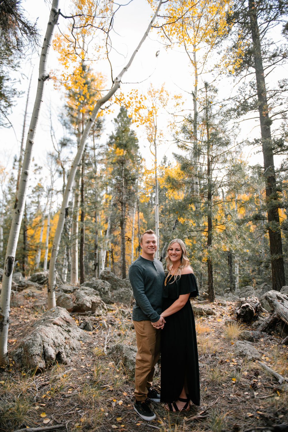 A couple stands together in a forest of aspen trees with golden autumn foliage and rocky terrain.