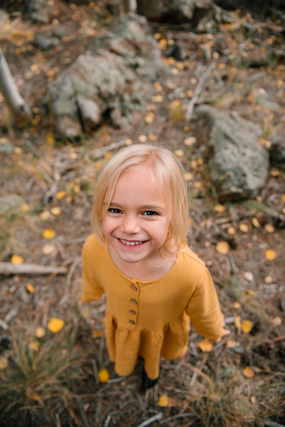 Child in mustard yellow dress smiling in autumn forest setting.