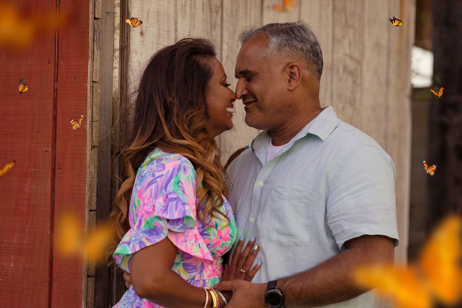 Happy couple sharing romantic moment nose-to-nose near rustic barn with orange butterfly bokeh background.