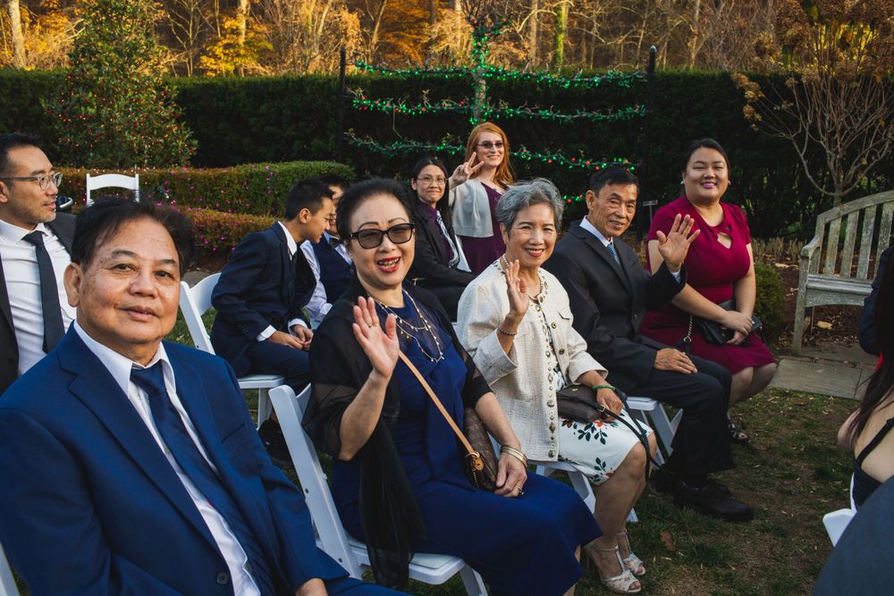 Wedding guests seated in formal attire wave to the camera during an outdoor garden ceremony.