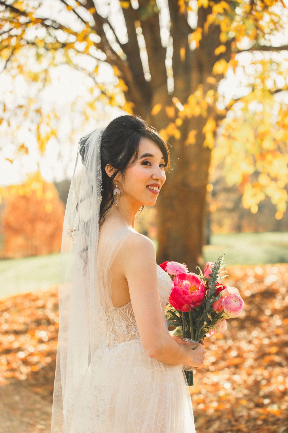 Bride holding bouquet of pink flowers while smiling and looking to the side at Brookside Garden in the fall foliage