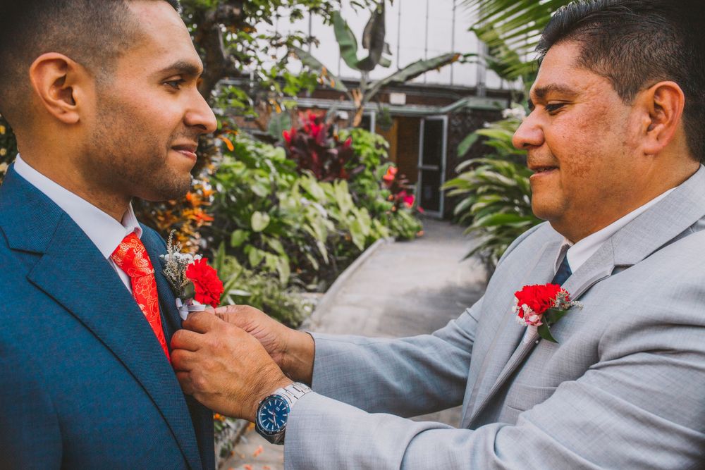 Two men in suits help each other with boutonnieres before a wedding ceremony.