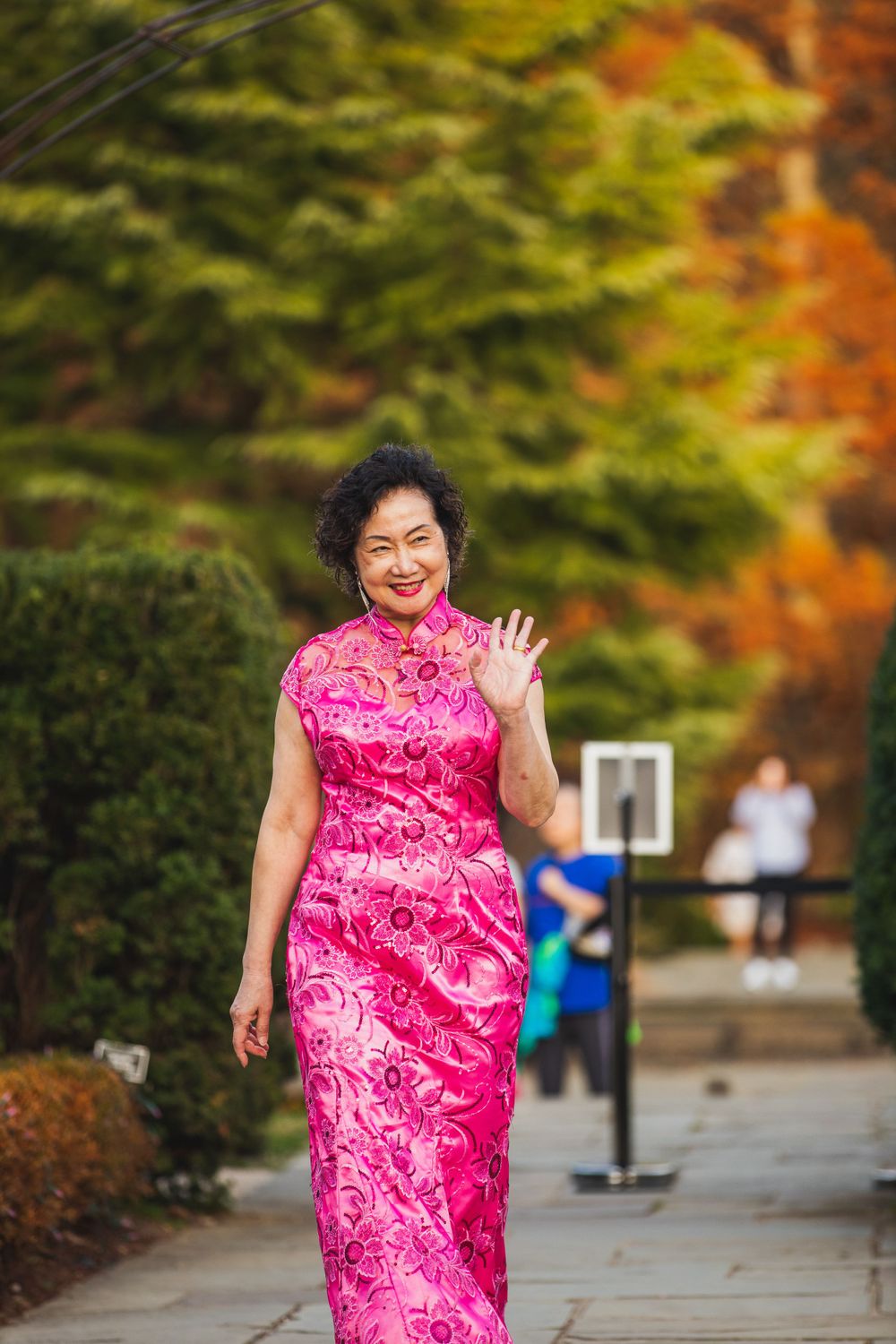 Woman in pink traditional dress waves while walking down garden path with fall colors behind.