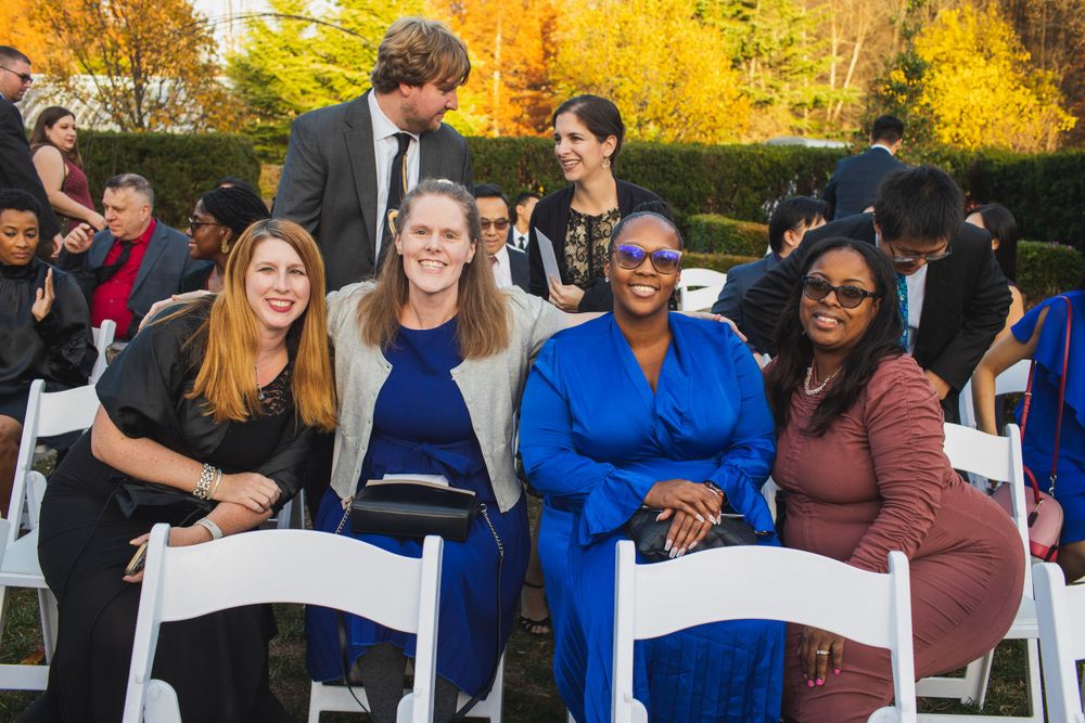 Wedding guests smile and laugh while seated in white chairs during an outdoor autumn ceremony.