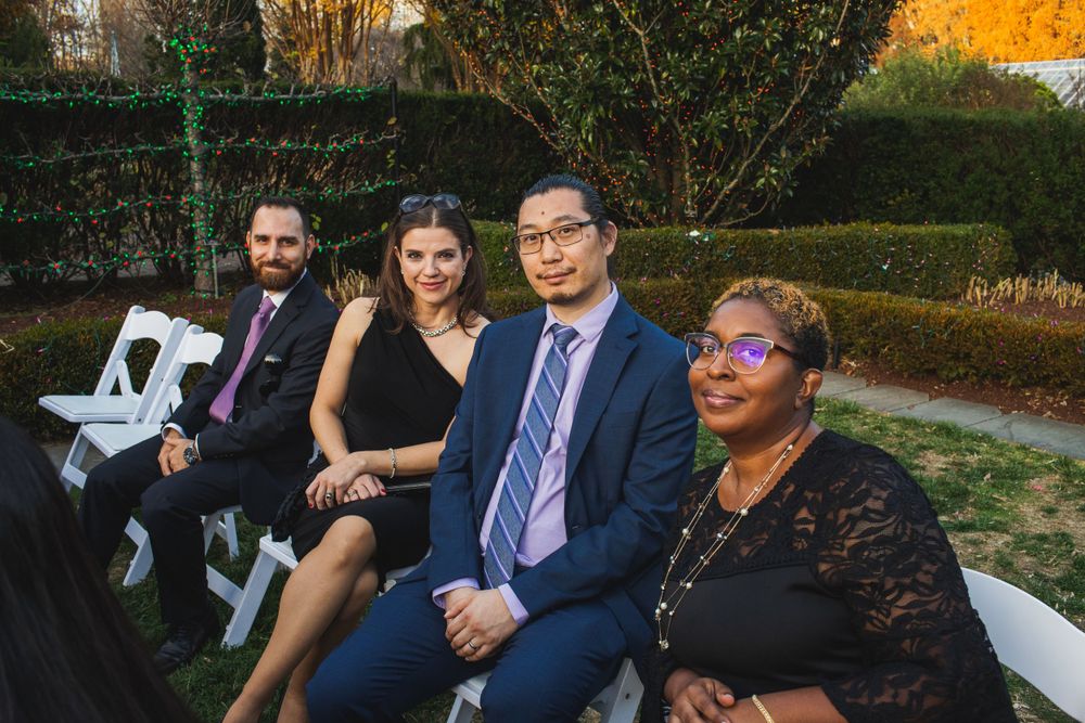 Group of wedding guests seated in white chairs at an outdoor garden event with fall foliage.