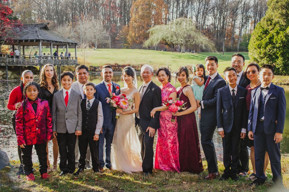 Large wedding party group photo with colorful attire against scenic fall backdrop with pond.