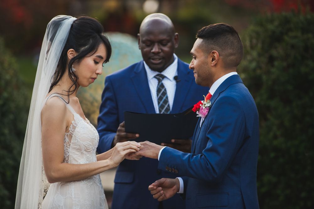 Couple exchanges rings during outdoor wedding ceremony with officiant standing between them.