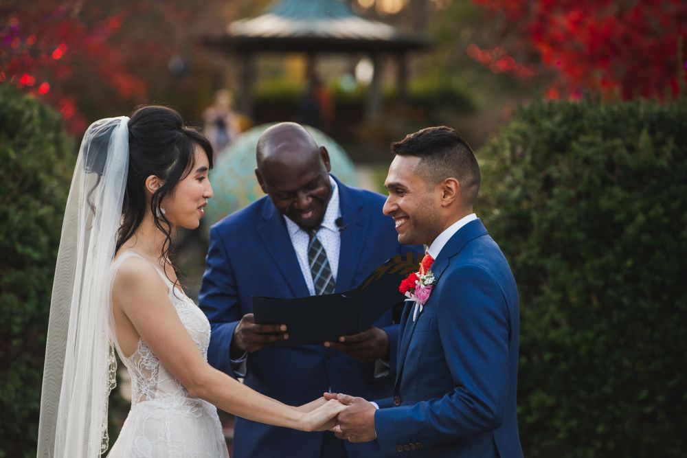 Bride and groom hold hands during wedding ceremony with officiant in blue suit.