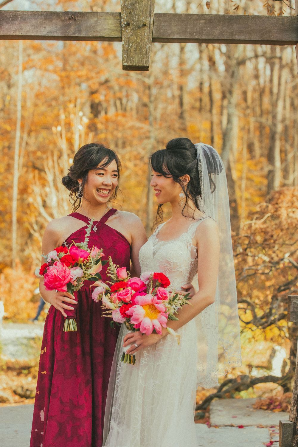 Bride and bridesmaid share joyful moment under wooden arch holding bright pink floral bouquets.