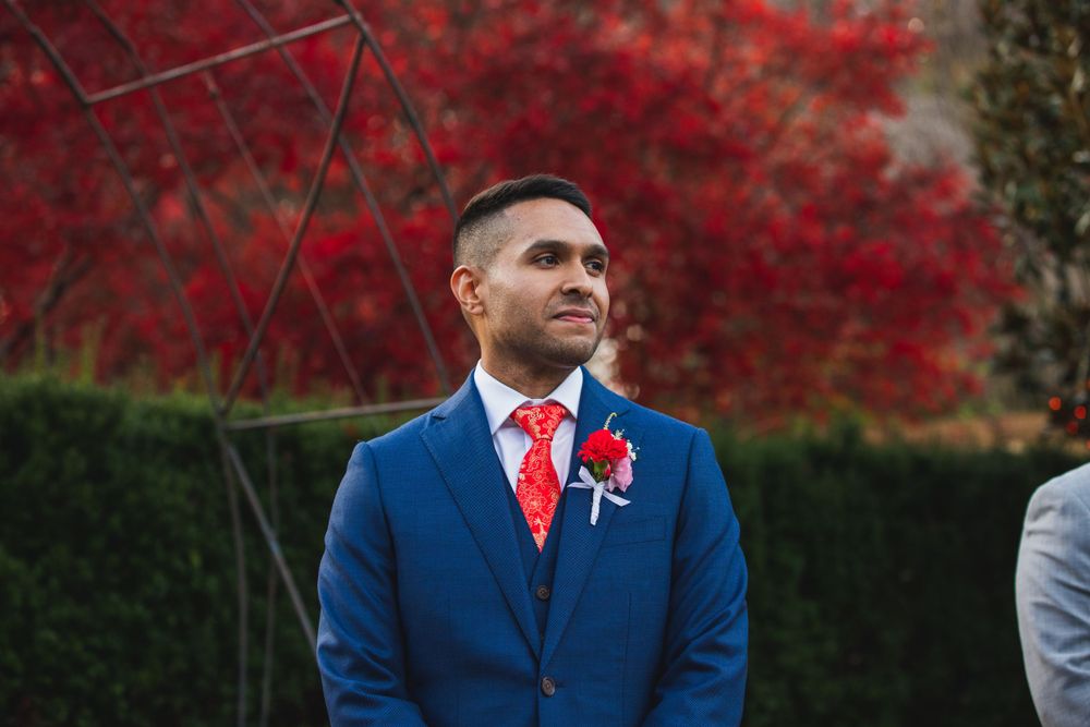 Groom in navy suit with red tie standing against vibrant autumn red maple backdrop.
