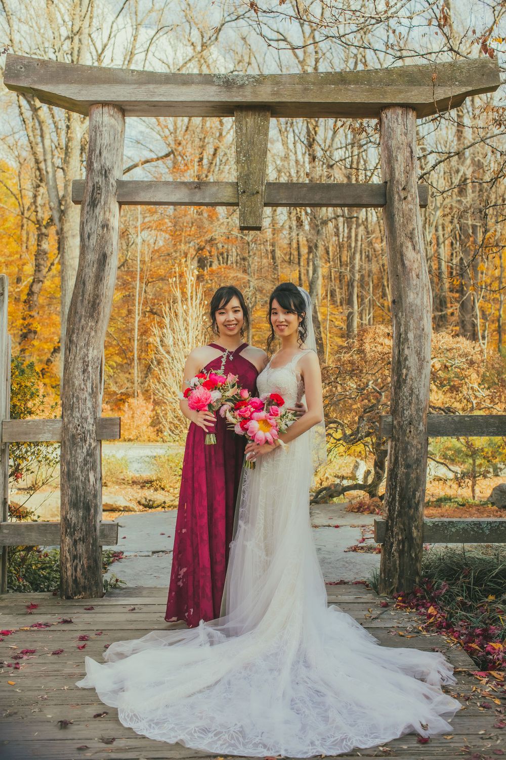 Wedding couple poses under rustic wooden arch surrounded by vibrant autumn foliage with burgundy and pink bouquets.