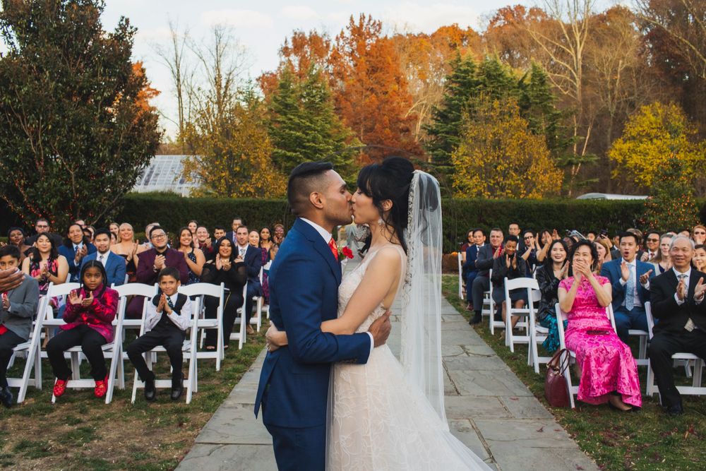 Newlyweds share first kiss at outdoor autumn wedding with guests seated on white chairs.