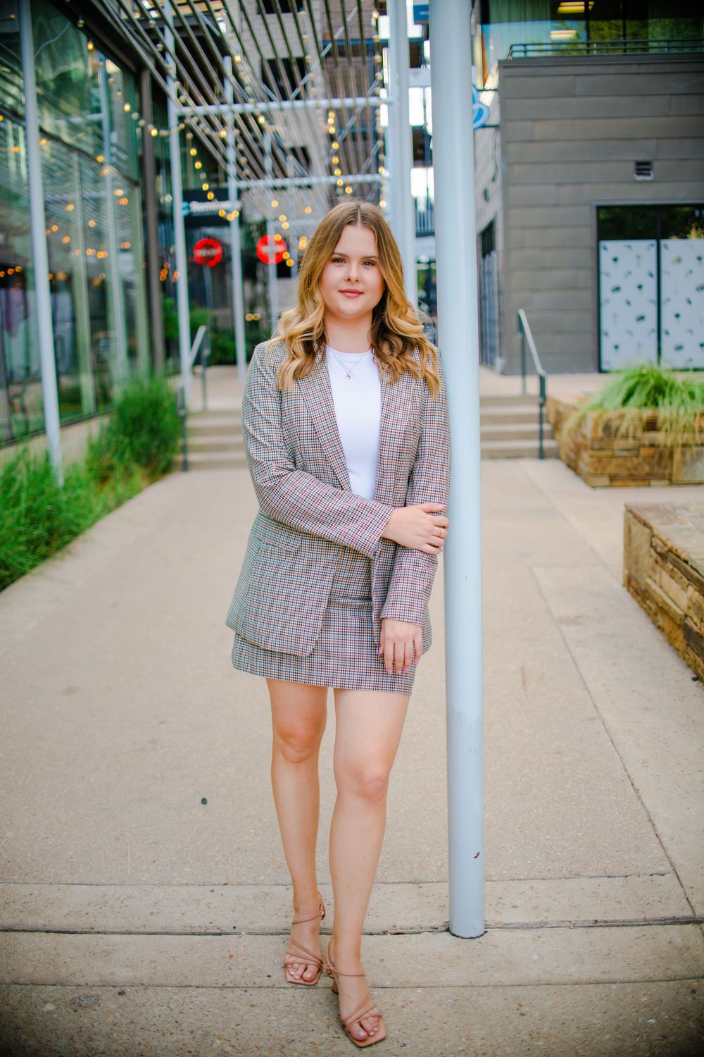 Person in plaid blazer poses on urban sidewalk near modern architecture.