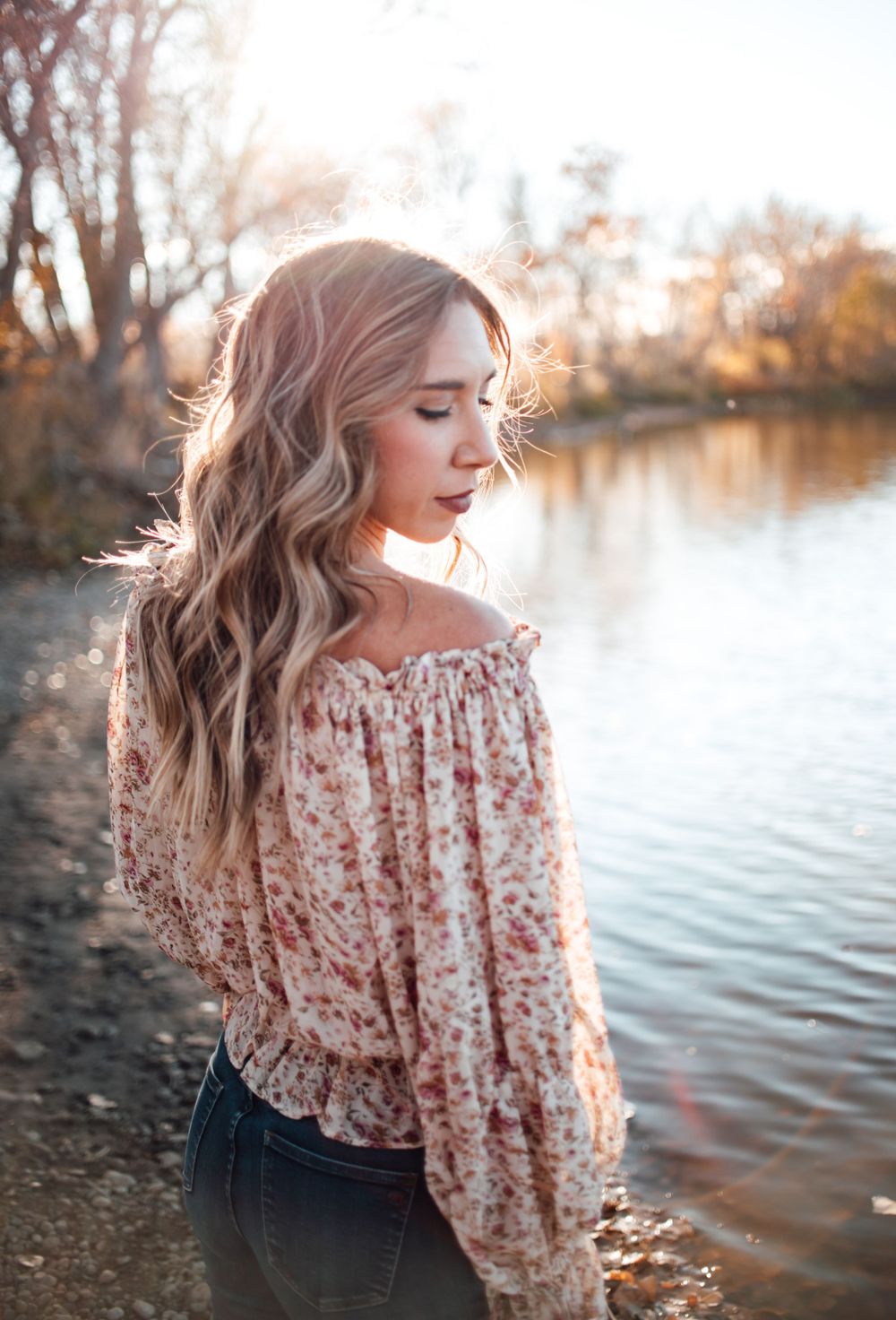 Woman in off-shoulder floral top poses by lake during golden hour portrait session.