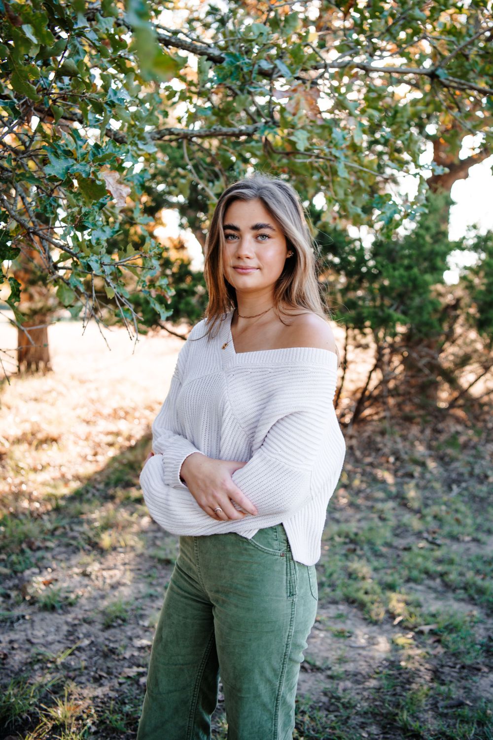 Person wearing white sweater and green pants poses outdoors among trees.