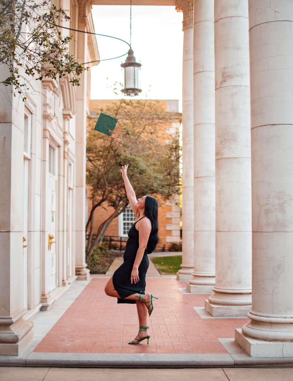 A person tosses a green graduation cap in a colonnade during a celebratory photoshoot.
