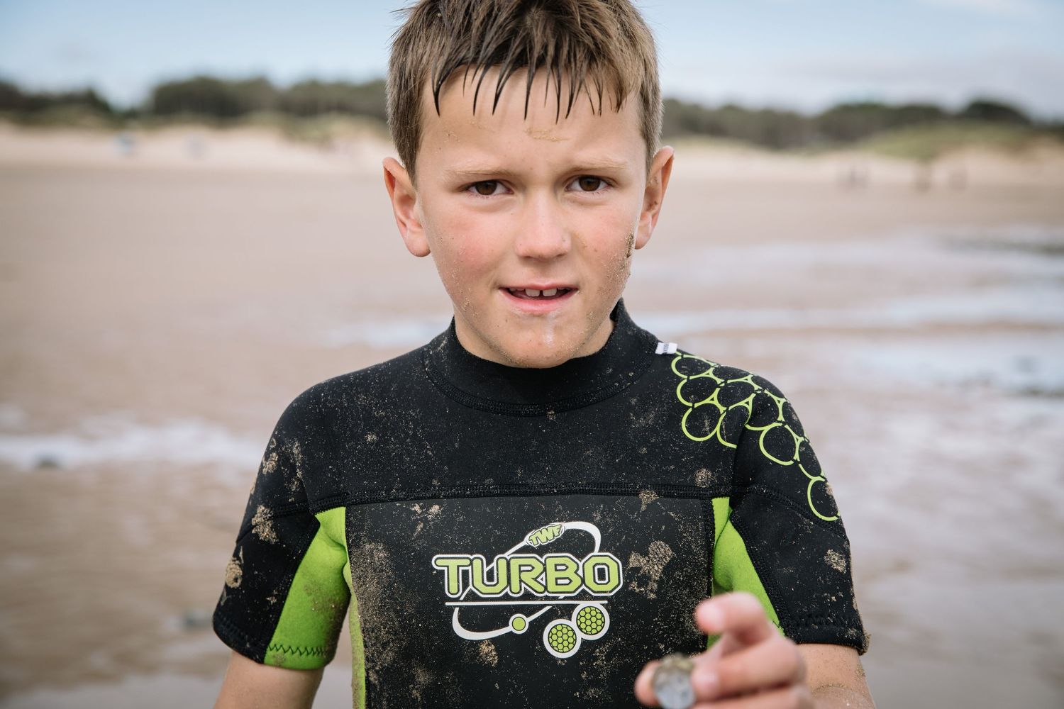 Child wearing a black and green Turbo wetsuit stands on a beach.