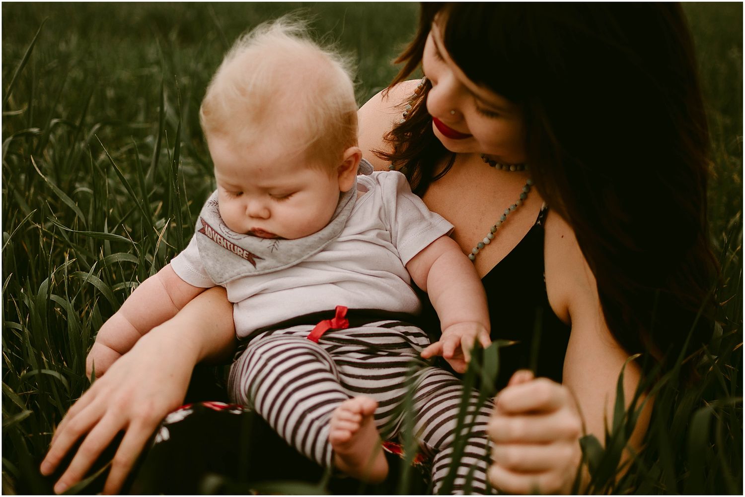 A baby in striped pants rests on a lap while sitting in tall grass outdoors.
