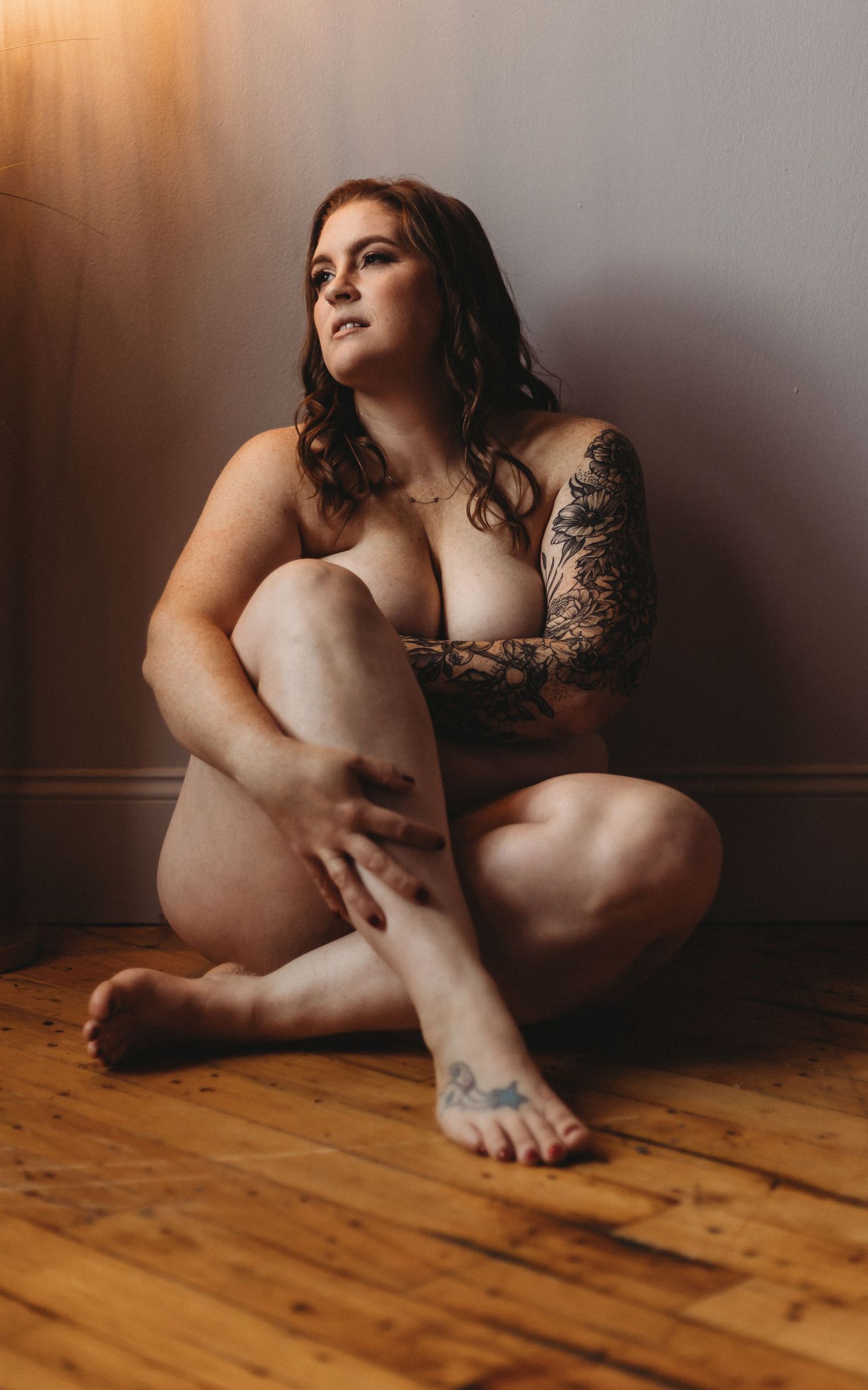 Artistic boudoir portrait of a body positive model with tattoos sitting on hardwood floor in moody lighting.