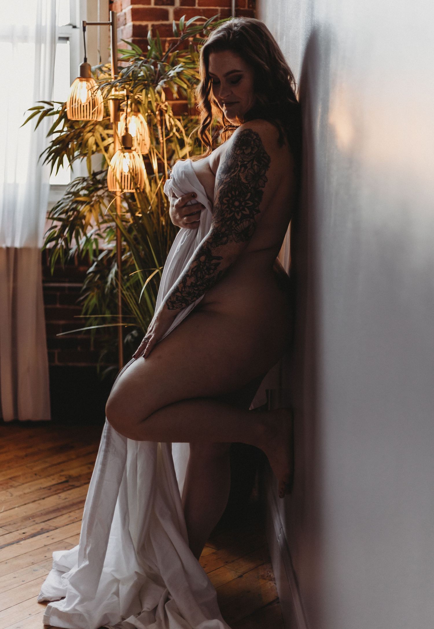Artistic boudoir photo with moody lighting features silhouette against exposed brick wall with hanging lights and plants.