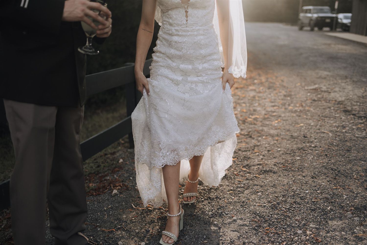 Detail shot of bride in lace dress walking with partner at sunset.