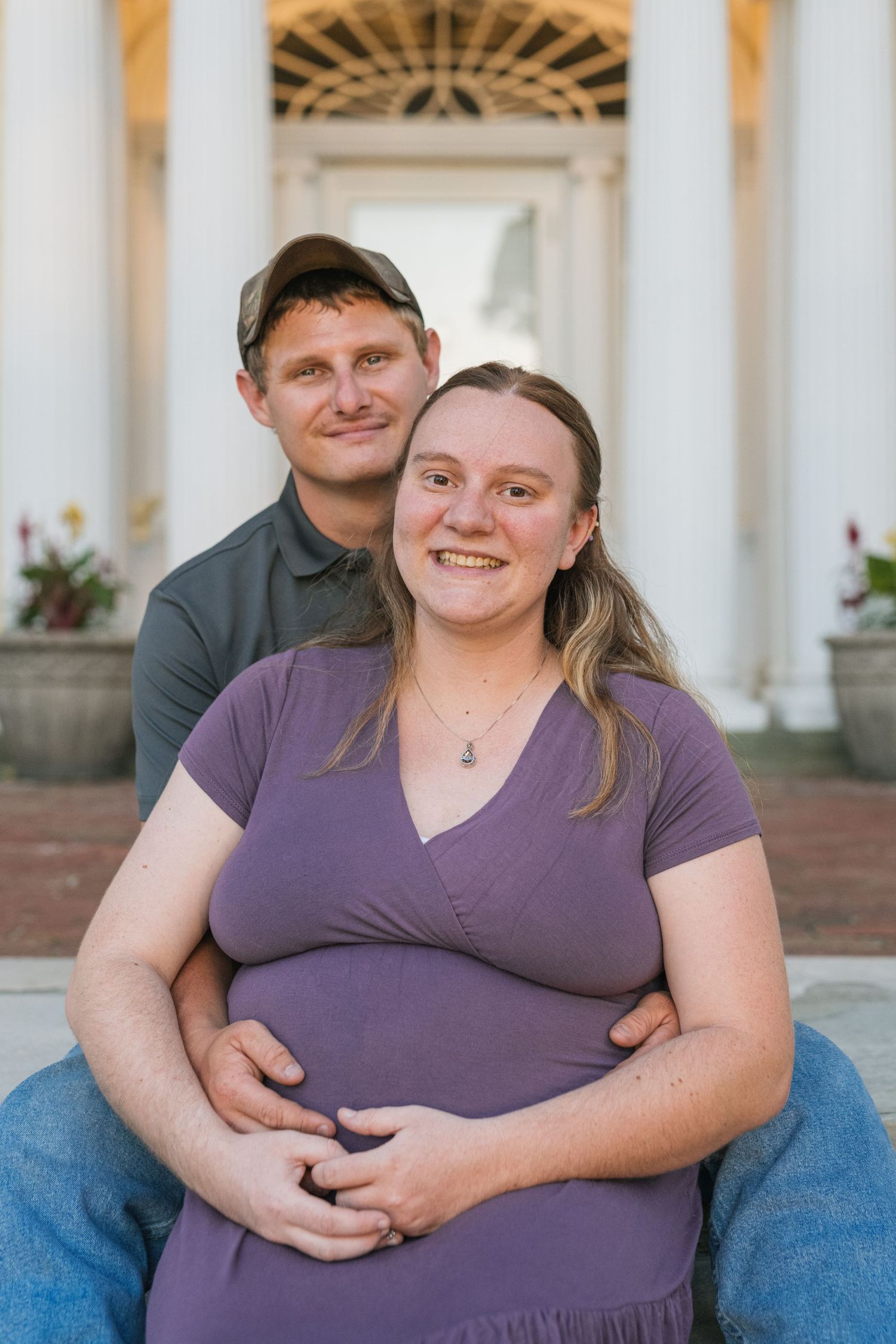 Couple embracing in a maternity pose with white pillars and ornate archway in the background.