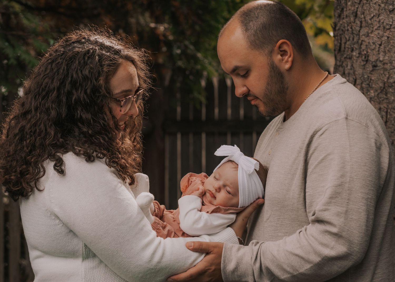A couple in white and beige sweaters tenderly hold their sleeping newborn baby outdoors near a tree in Uxbridge, Massachusetts.