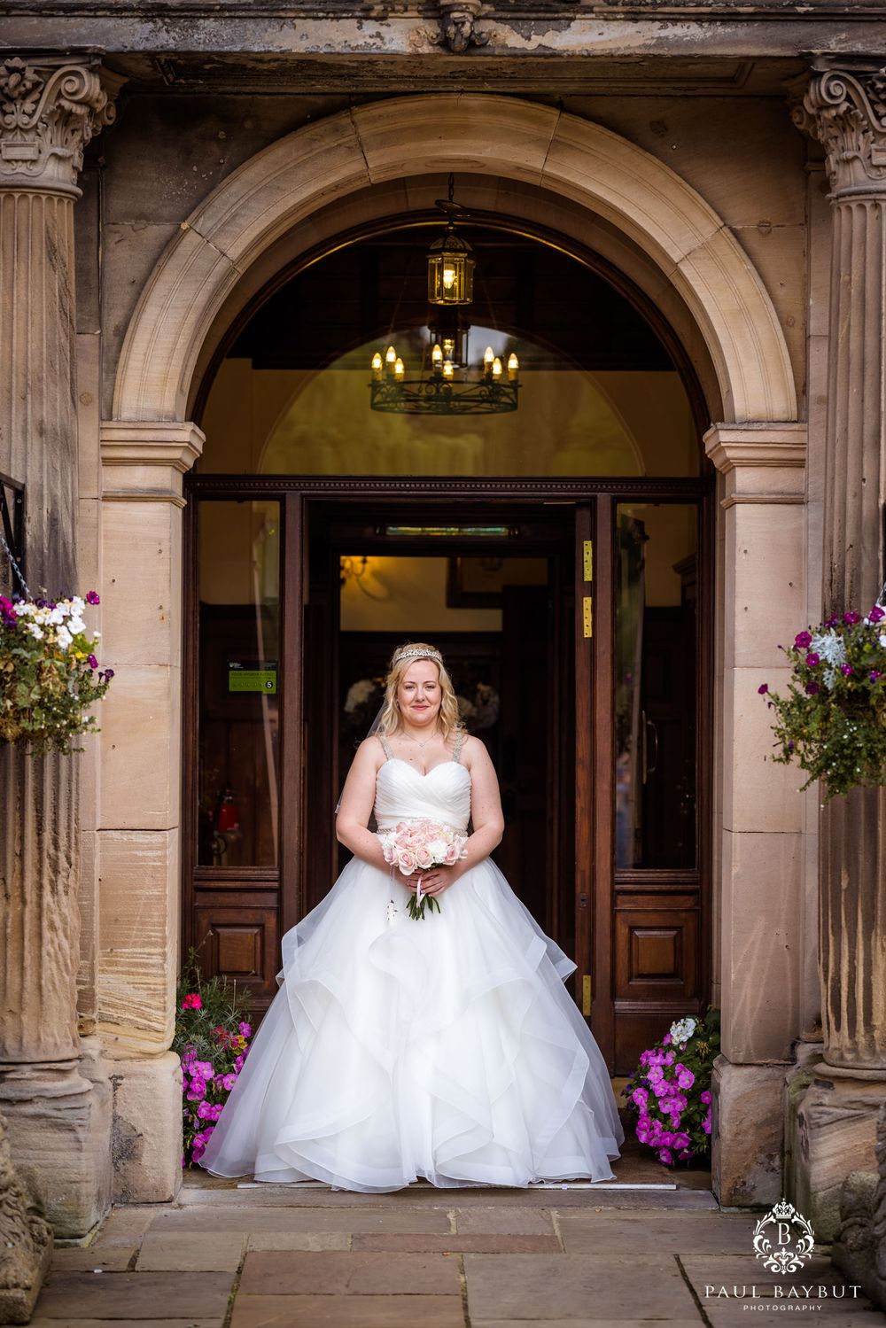 Elegant bride in full ball gown stands under stone archway adorned with purple flowers.