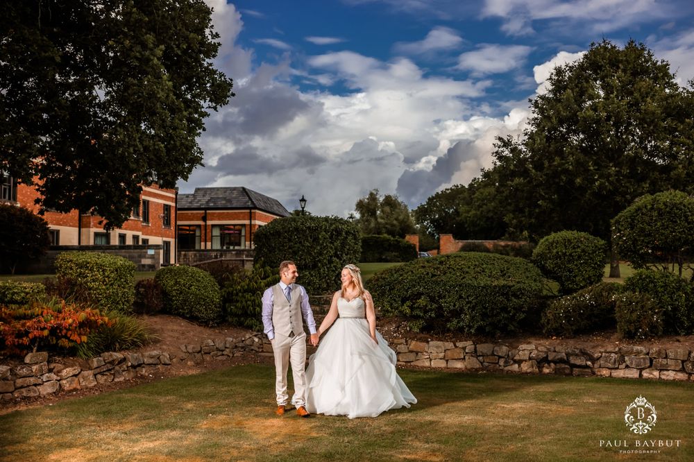 Wedding couple poses on manicured grounds with dramatic sky and brick building backdrop.