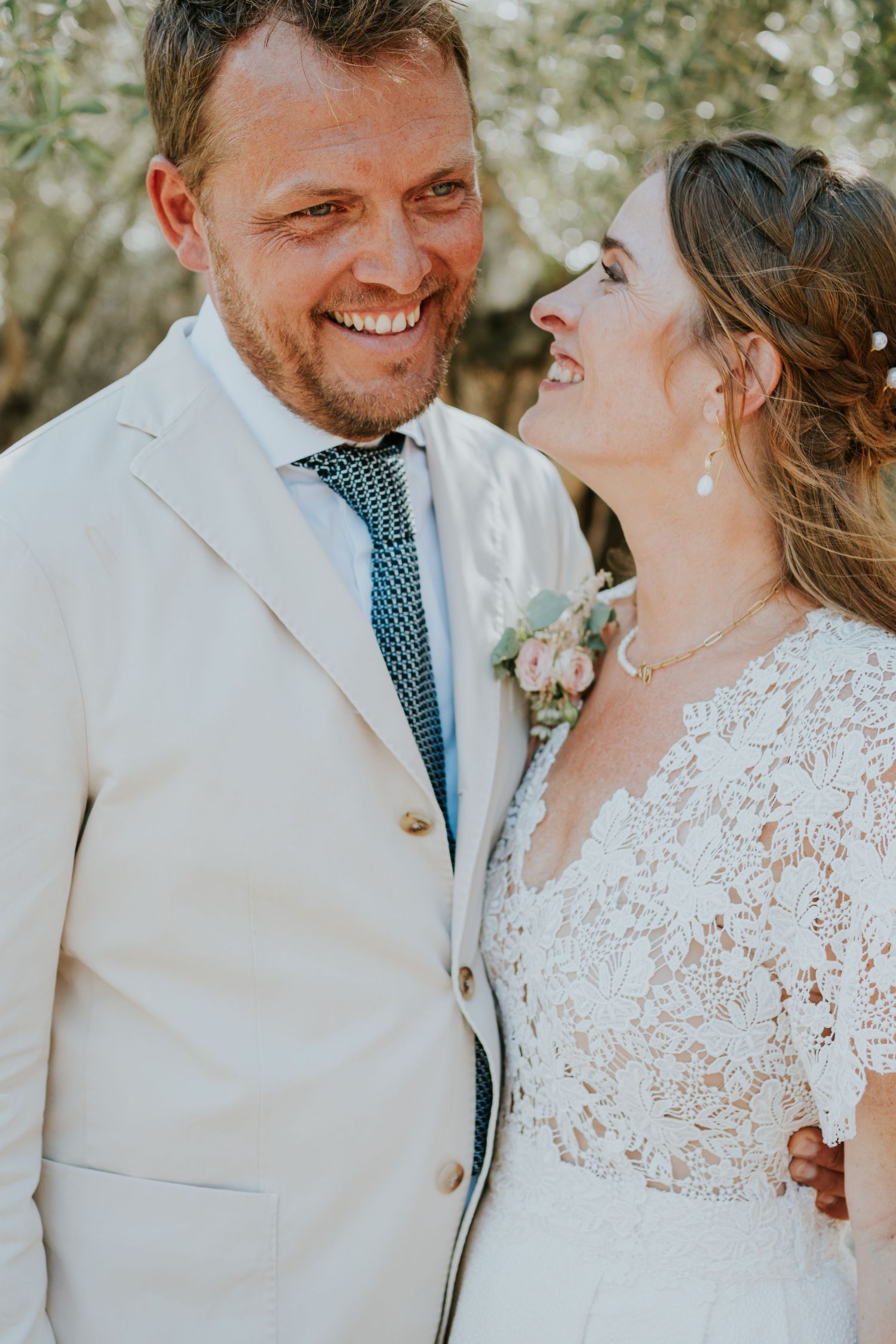 A romantic wedding couple shares a joyful moment in their lace and cream-colored attire against a natural backdrop.