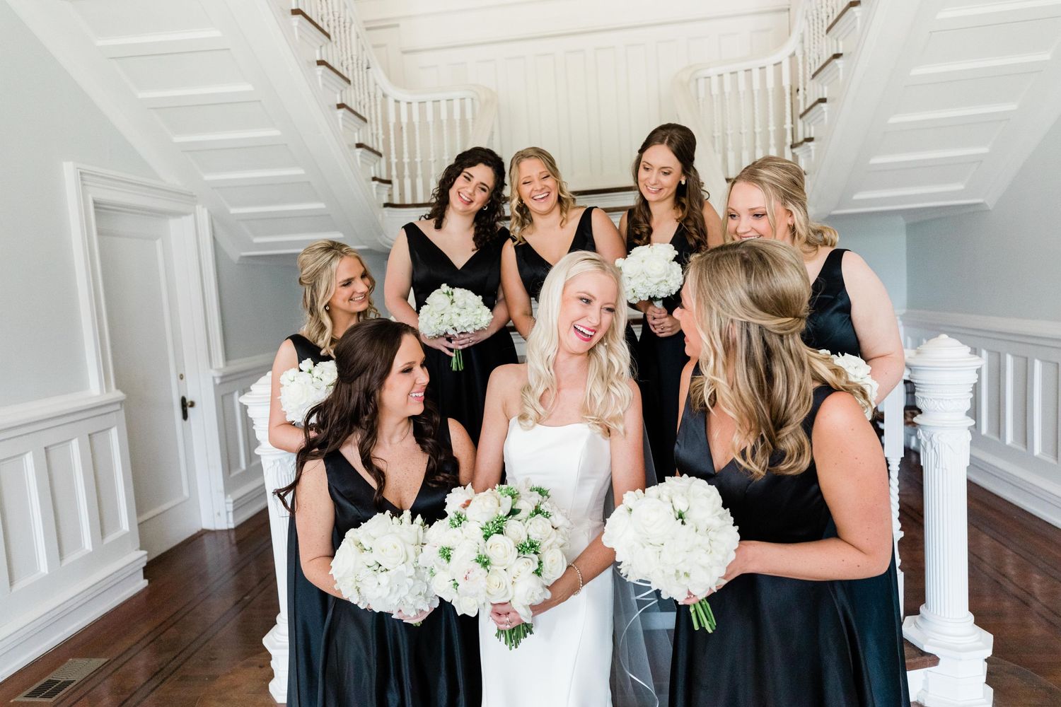 Bridesmaids in black dresses gather for indoor photo with white floral bouquets.