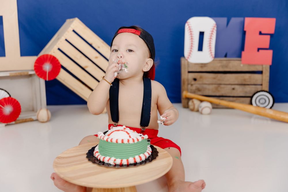 A baby boy is eating a cake in front of a baseball field.