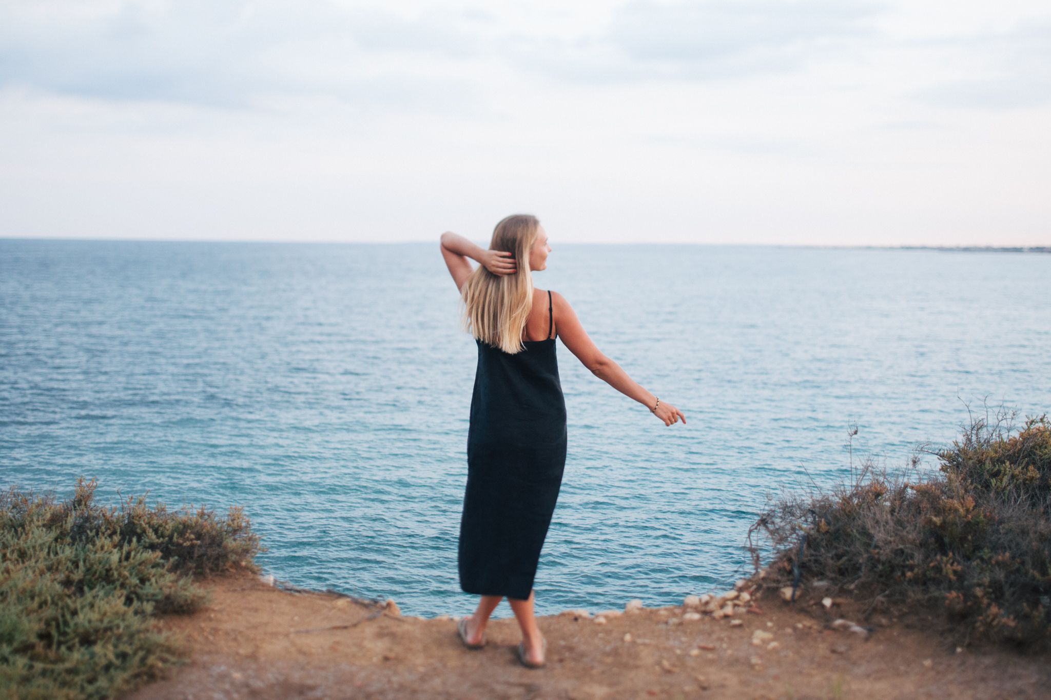 Blonde woman in black dress standing on rocky cliff edge overlooking calm blue ocean.