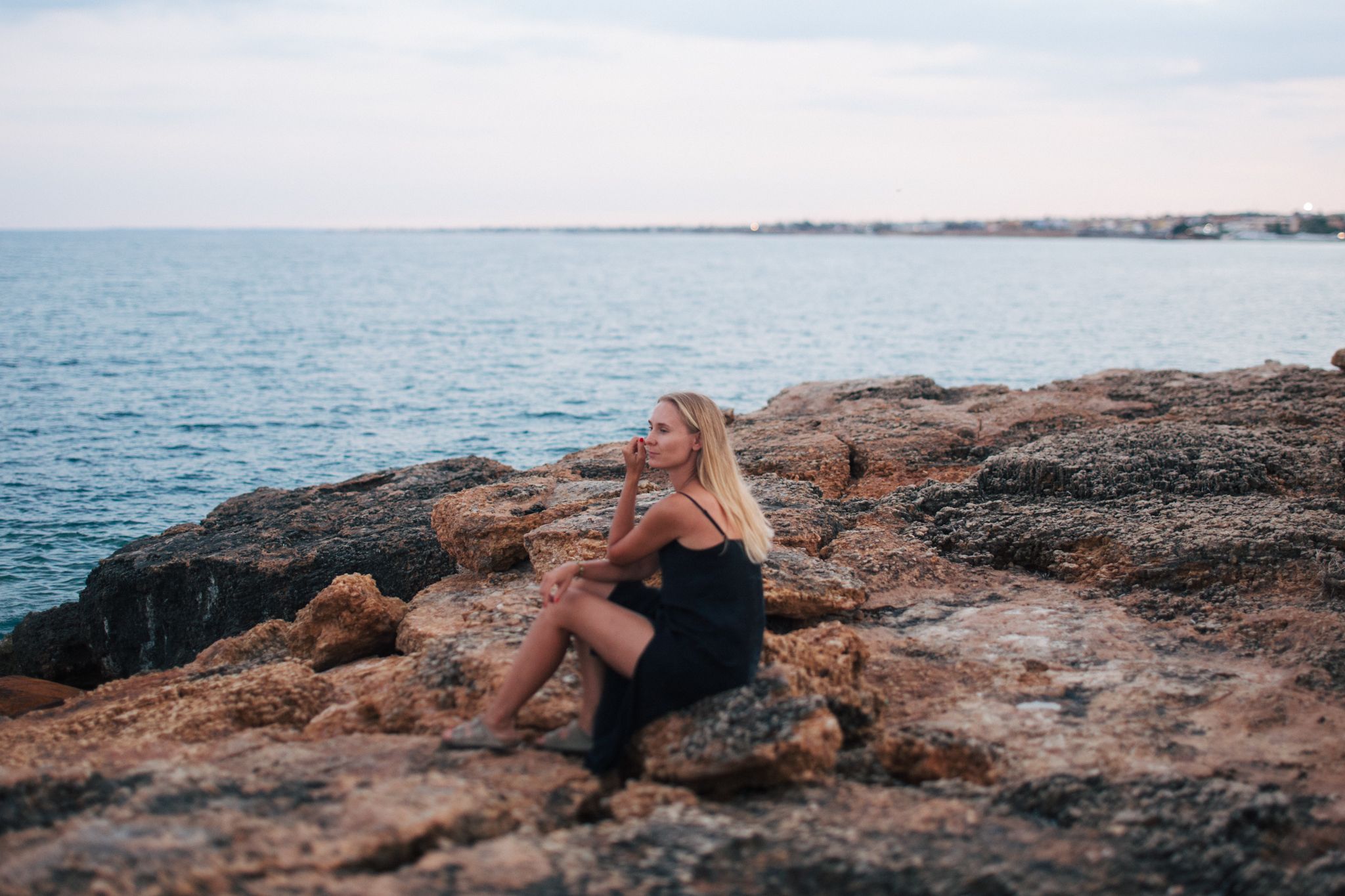 Blonde woman in black dress sitting on rocky coastal shore gazing at the sea.