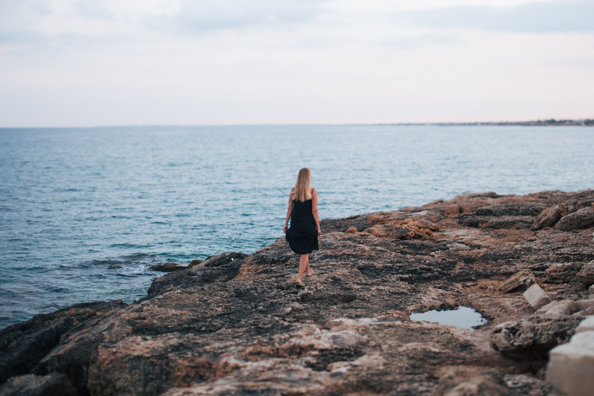 Blonde woman in black dress standing on rocky coastline looking out at the ocean.