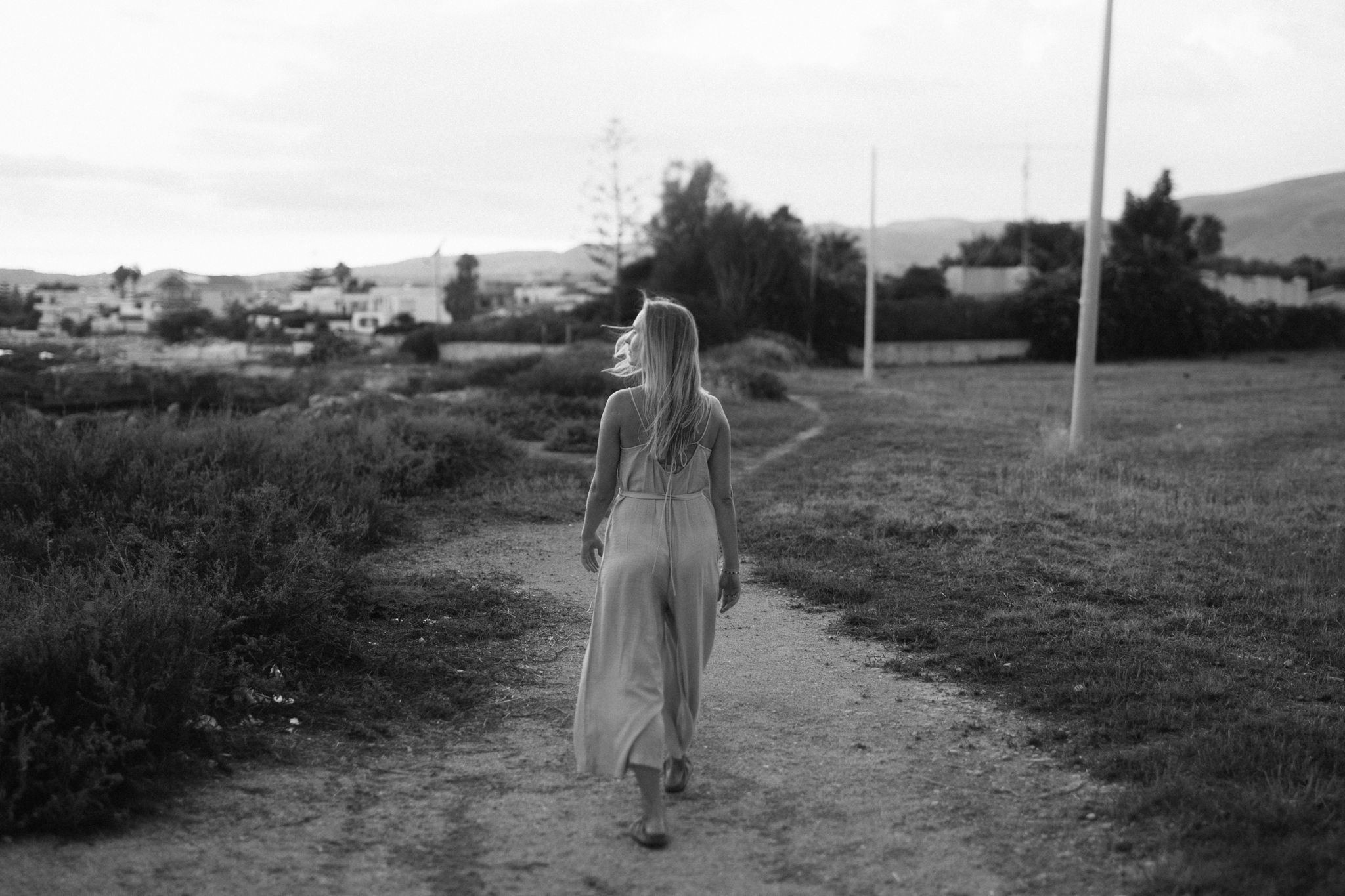 Blonde woman in white dress walking along a rural dirt path in black and white photo.