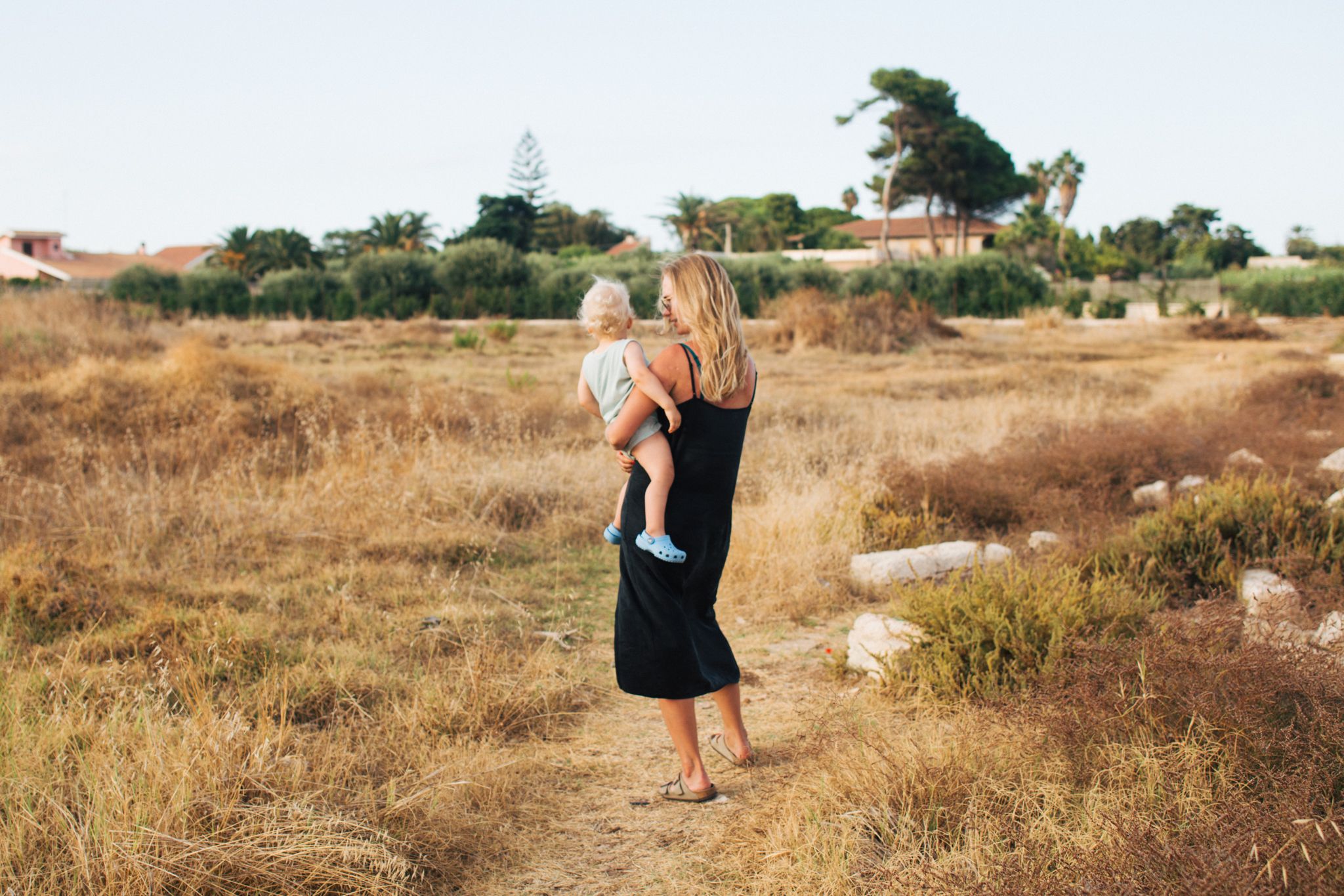 Blonde mother in black dress holding baby while walking through dry golden field.