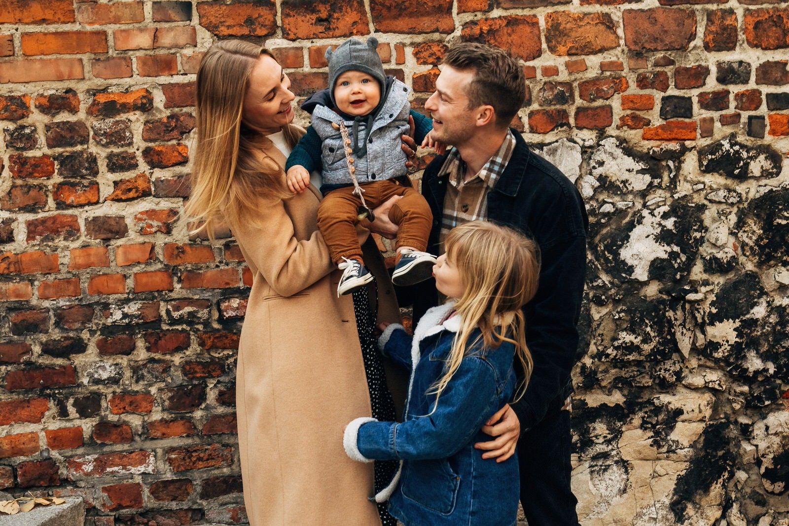 Smiling family of four posing together against rustic brick wall in autumn outfits.