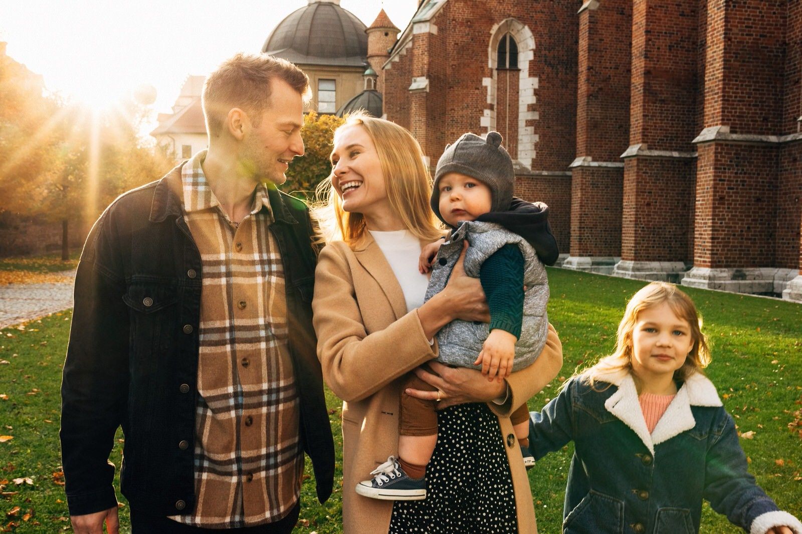 Happy young family with two children smiling outdoors near brick building in autumn sunlight.