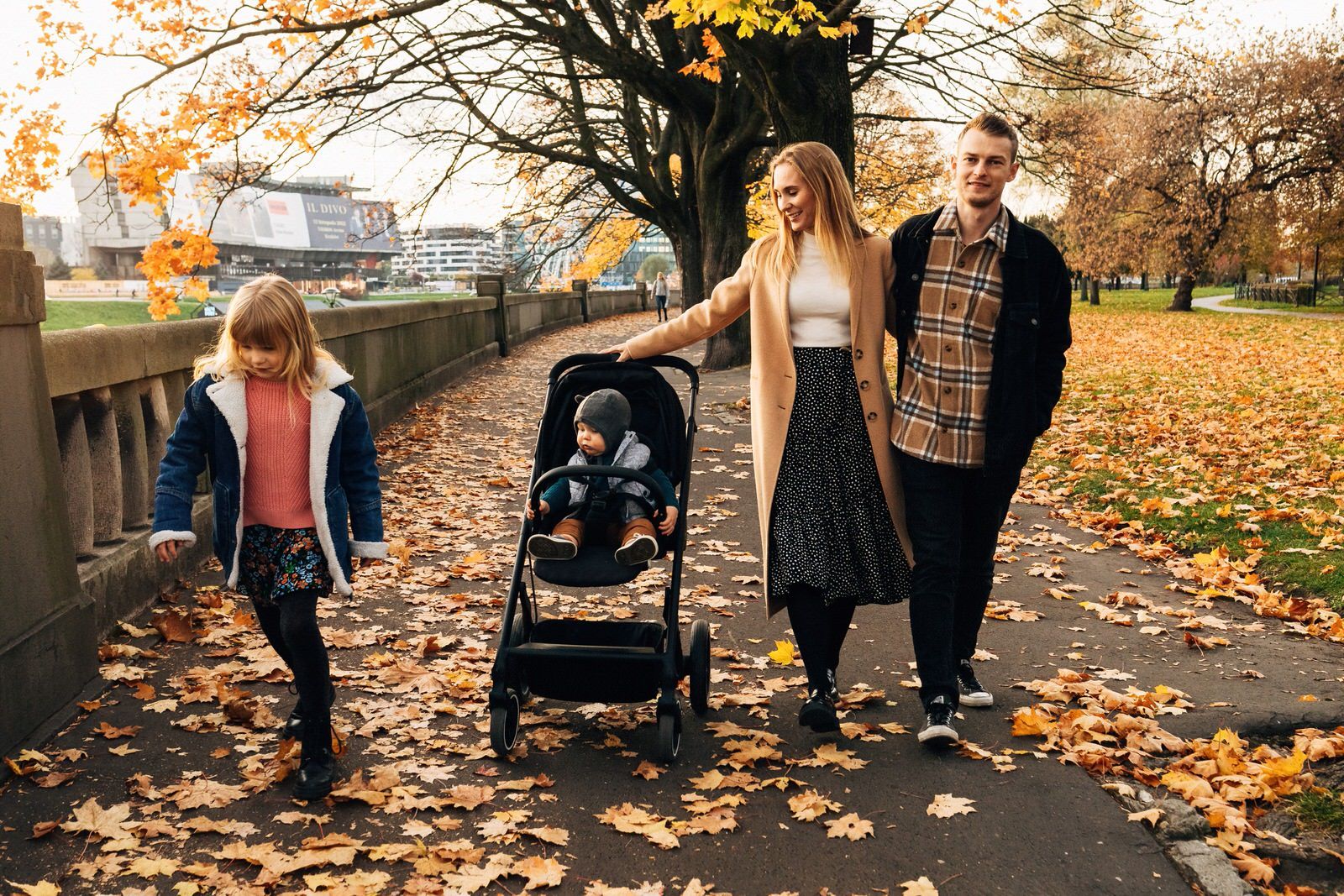 Young family strolling through autumn park with baby stroller and fallen leaves on path.