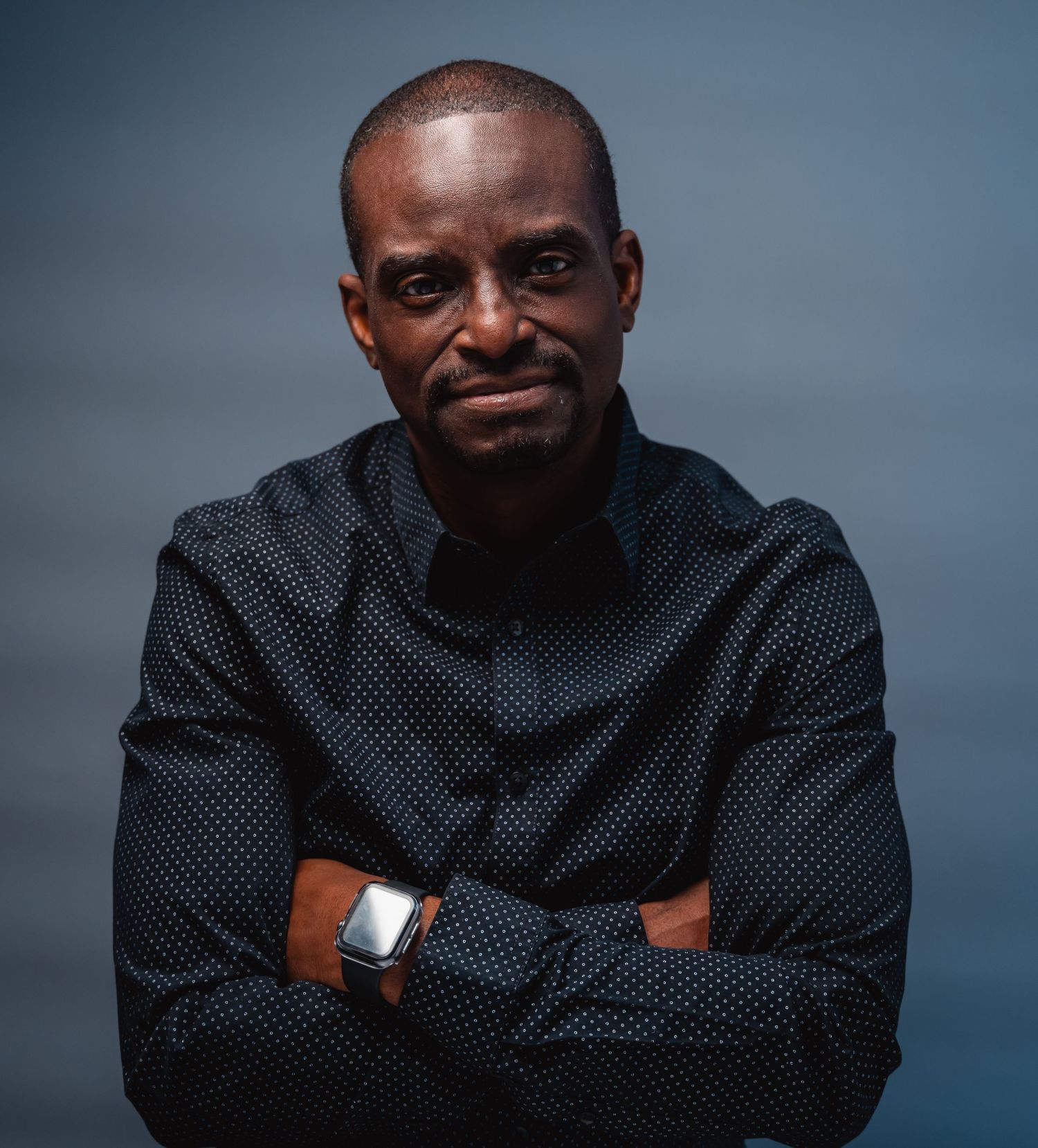 Man in black dotted shirt standing with arms crossed confidently.