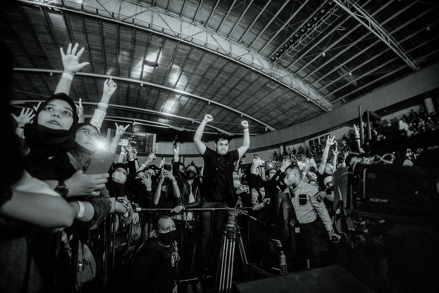 Black and white shot of concert crowd with raised hands under industrial ceiling.